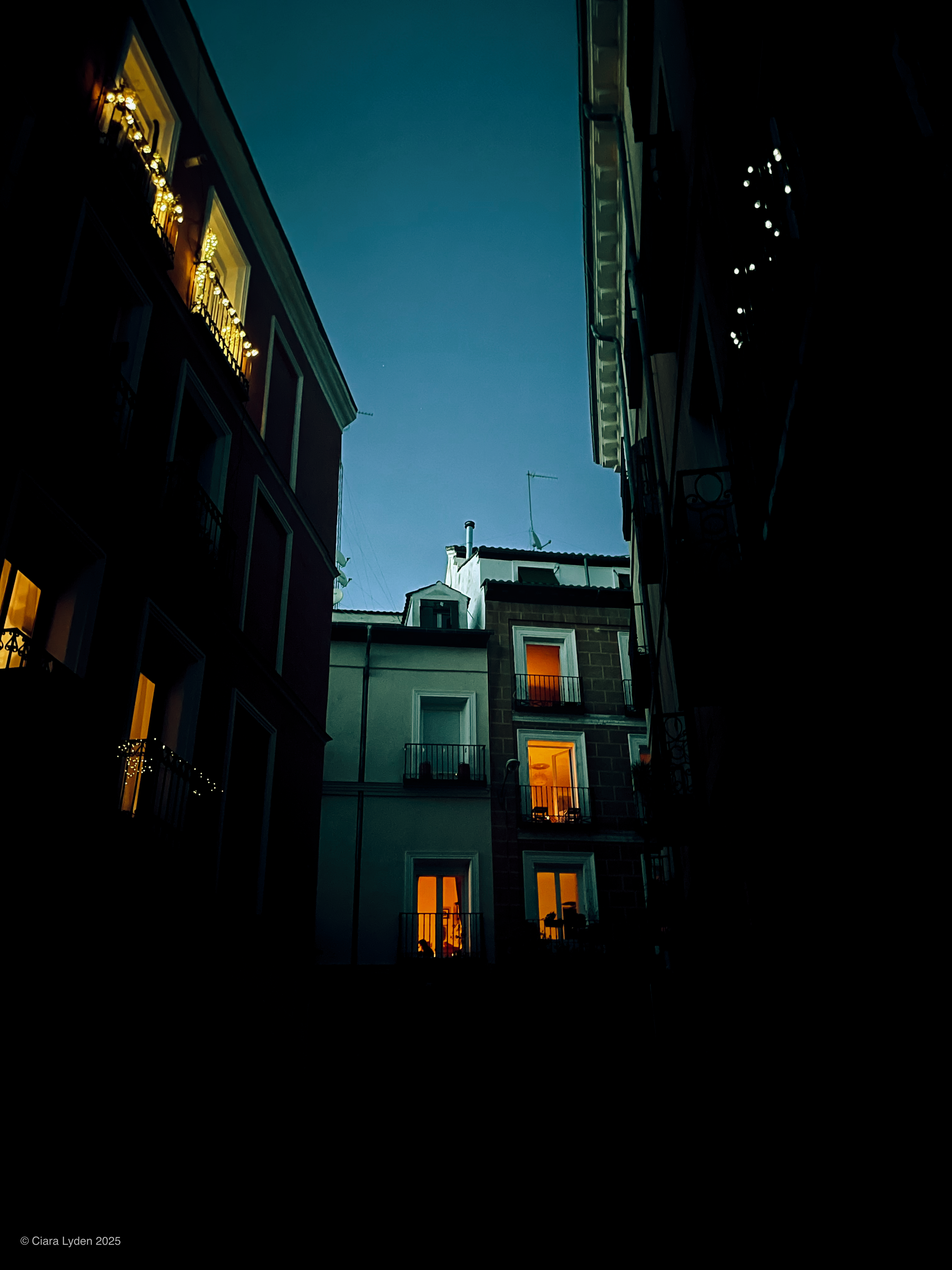 Looking up between Madrid apartment blocks at dusk. Eight windows in the buildings are lit warm orange from candles inside. Another window on the left is faintly lit. String lights hang from a balcony at the top left. The sky is pale fading blue.