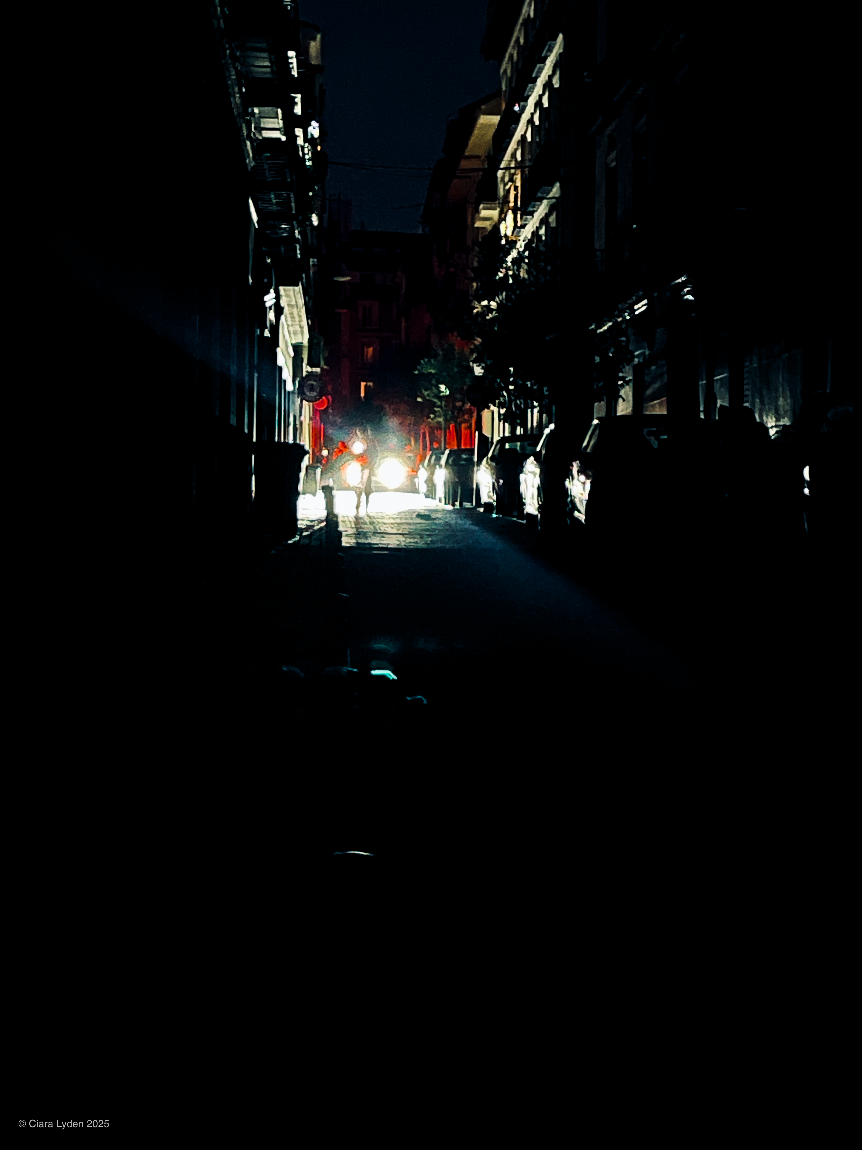 A narrow Madrid street at night. Two bright white headlights at the far end illuminate silhouetted figures around them. The buildings on either side are completely dark.