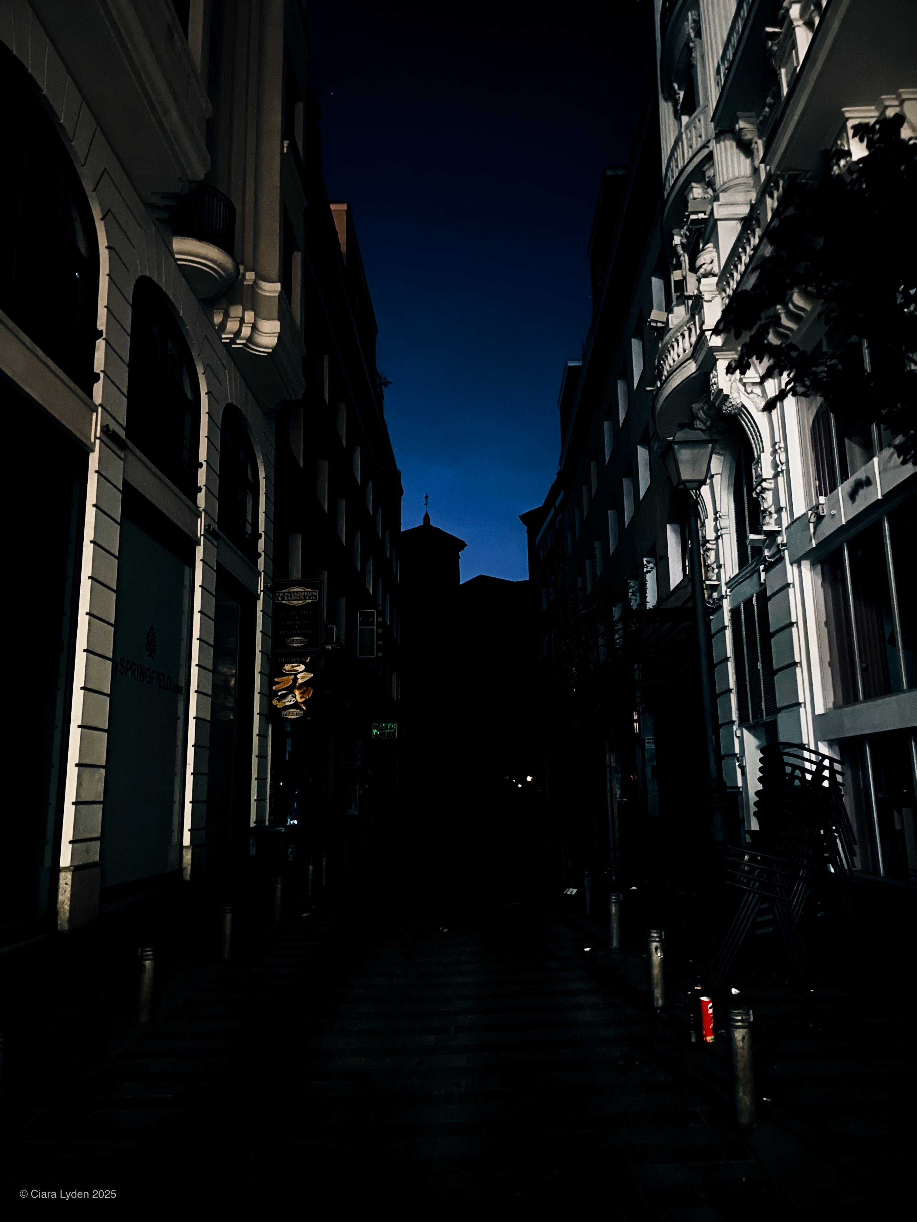 A Madrid shopping street at night, viewed from one end. The storefronts are dark and unlit. The sky between the buildings is a deep saturated blue.