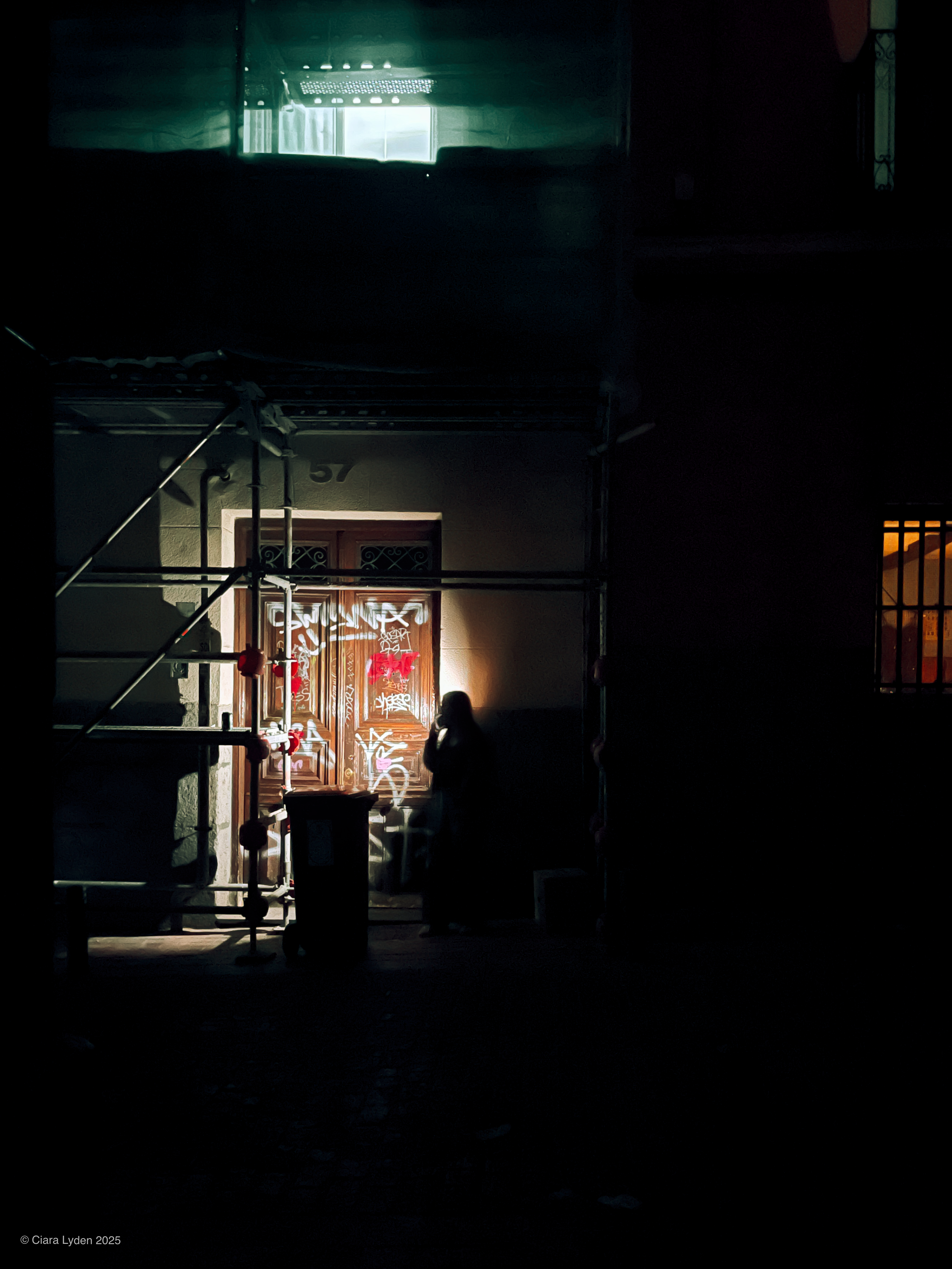 A doorway on a Madrid street at night. The door is heavily covered in graffiti, visible in the orb of light cast from a figure standing in front of it. They are on their phone with the torch illuminated, lighting them from behind; they are otherwise mostly in shadow, a silhouette. Scaffolding frames the left side. A faint blue-green light spills from a window above. A warm orange light glows from a window on the right. The image was taken after the city had begun restoring energy but before the street lights came back on.