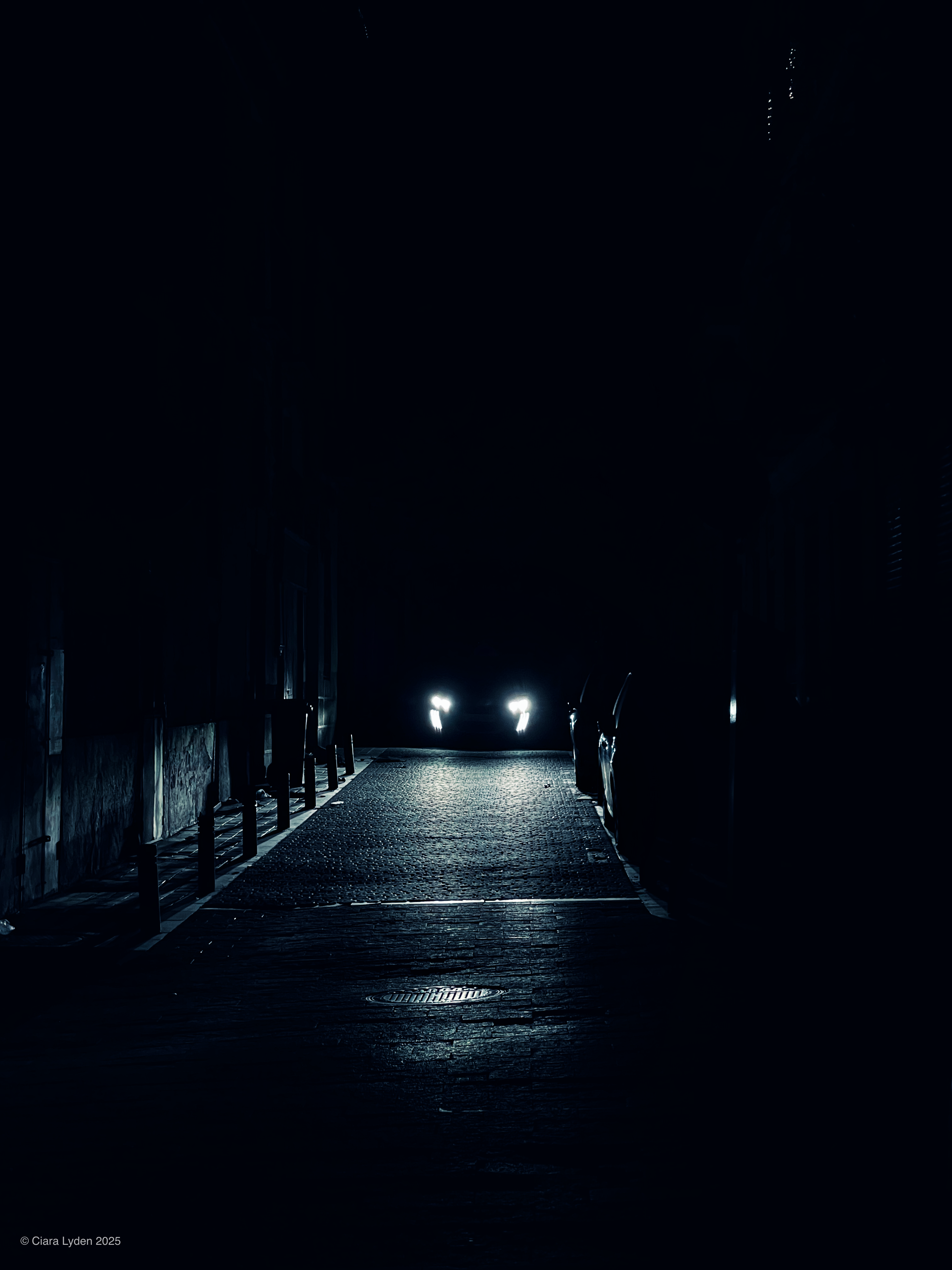 A dark alley at night. A single car at the far end has its headlights on, casting a narrow beam of light along the cobblestones toward the camera. The walls and surroundings are entirely black.