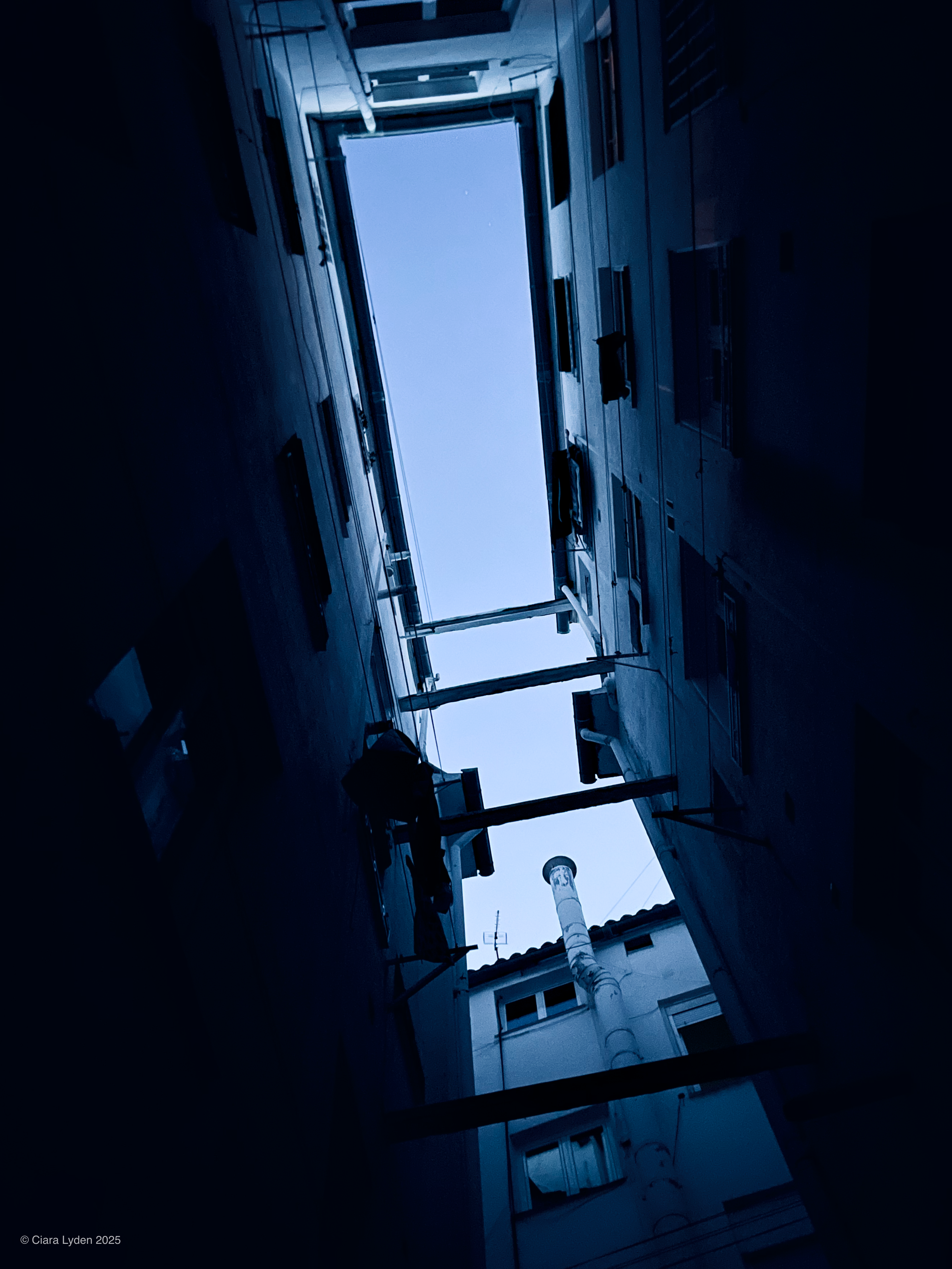 A vertical view looking up between two narrow Madrid apartment buildings at dusk. A pale blue sky shows at the top. A white drainpipe and a small antenna are silhouetted against the sky. The walls of the buildings are dark. Windows are visible and not one window has a light on.