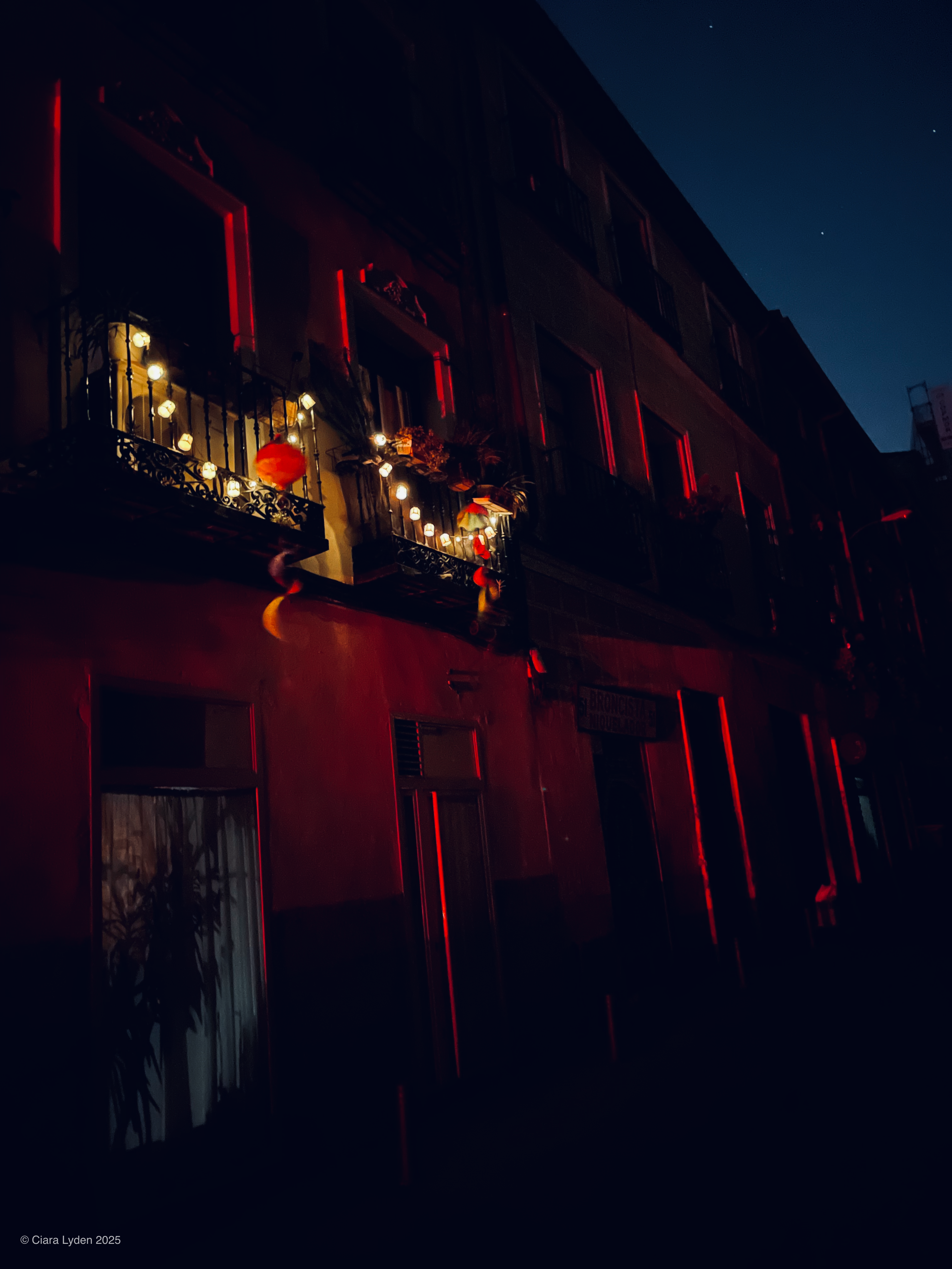 A Madrid building facade at night. A balcony in the upper centre is strung with paper lanterns and small lights. Red light from a nearby ground-level source falls across the stucco wall and the balcony railings. The sky behind is deep blue at the edge of night.