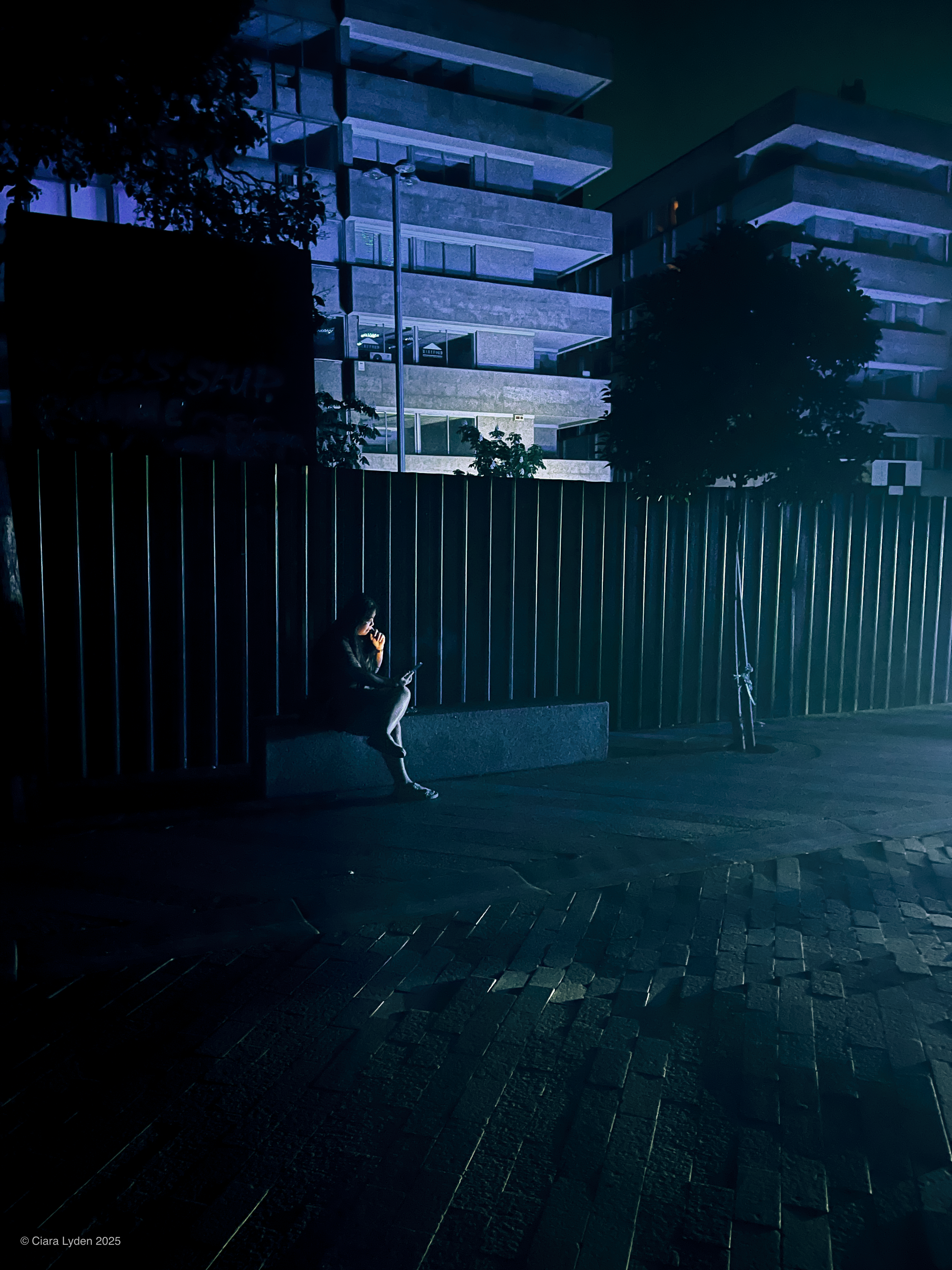 A person sits alone on a low concrete bench at night, lit only by the screen of their phone. Behind them, a tall modern office block is lit in cold blue tones. A metal fence runs between the bench and the building.