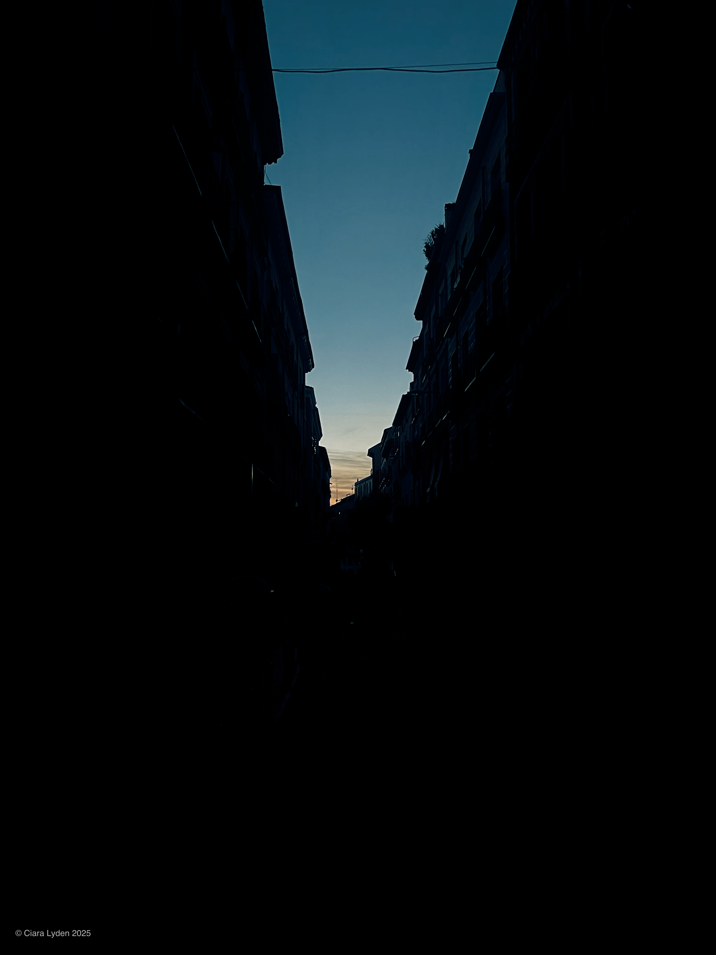Looking down a narrow Madrid street at twilight. The buildings on both sides are pure silhouette. A small wedge of orange and pale yellow sunset shows at the end of the street. A power line crosses the sky.