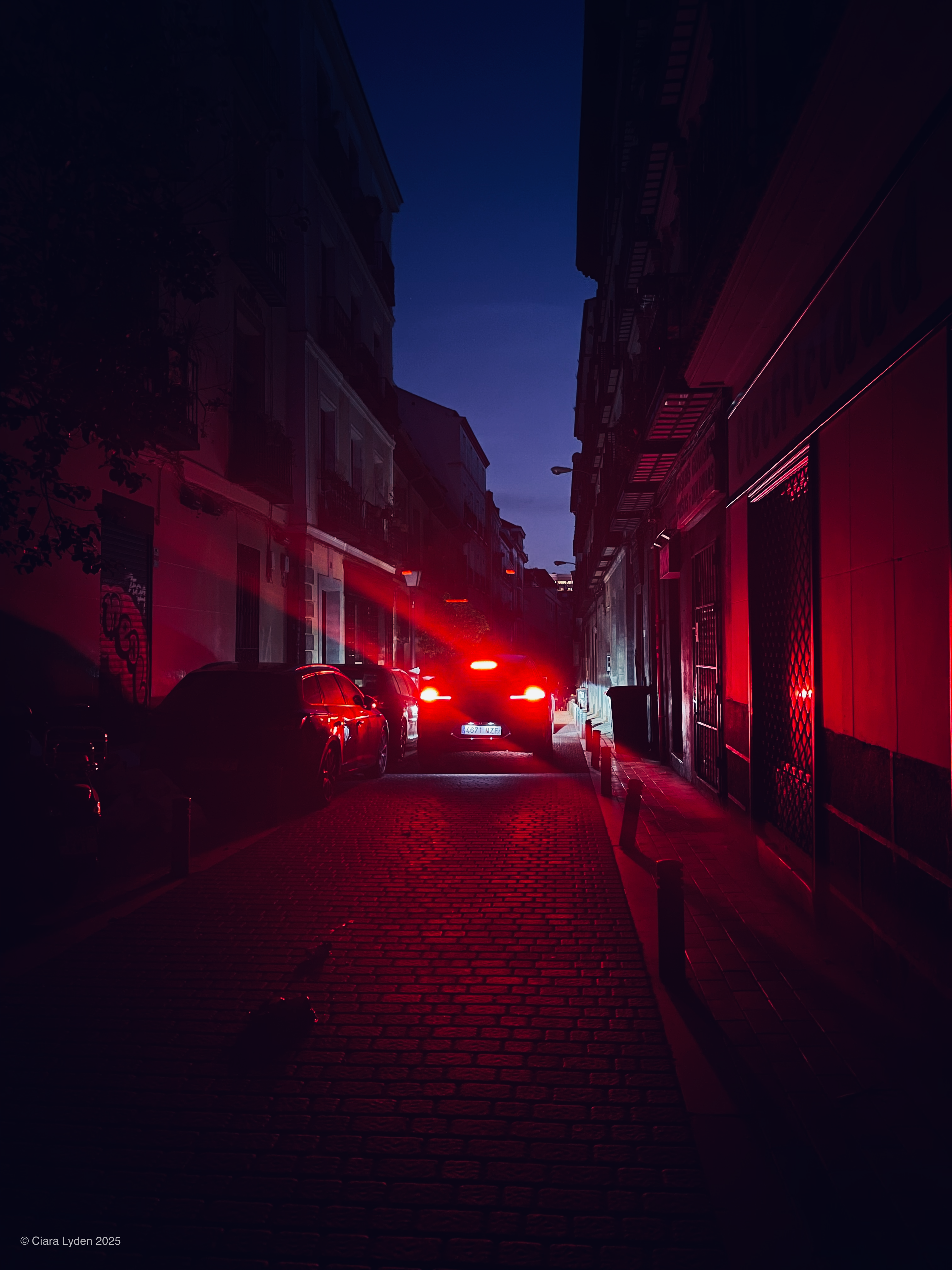A narrow Madrid street at night. The red brake lights of a car in the centre of the street wash the entire scene in red. Parked cars line the left side. The sky above is deep blue.